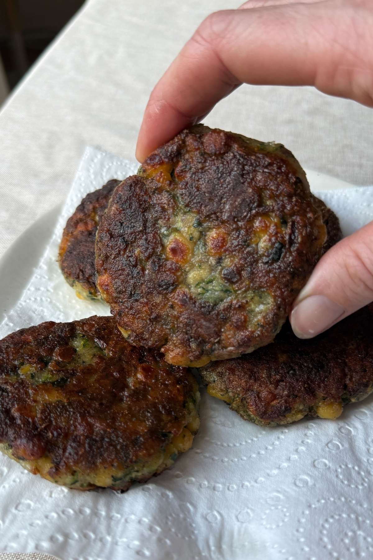 Hand holding a crispy zucchini chickpea fritter over a paper towel-lined plate.