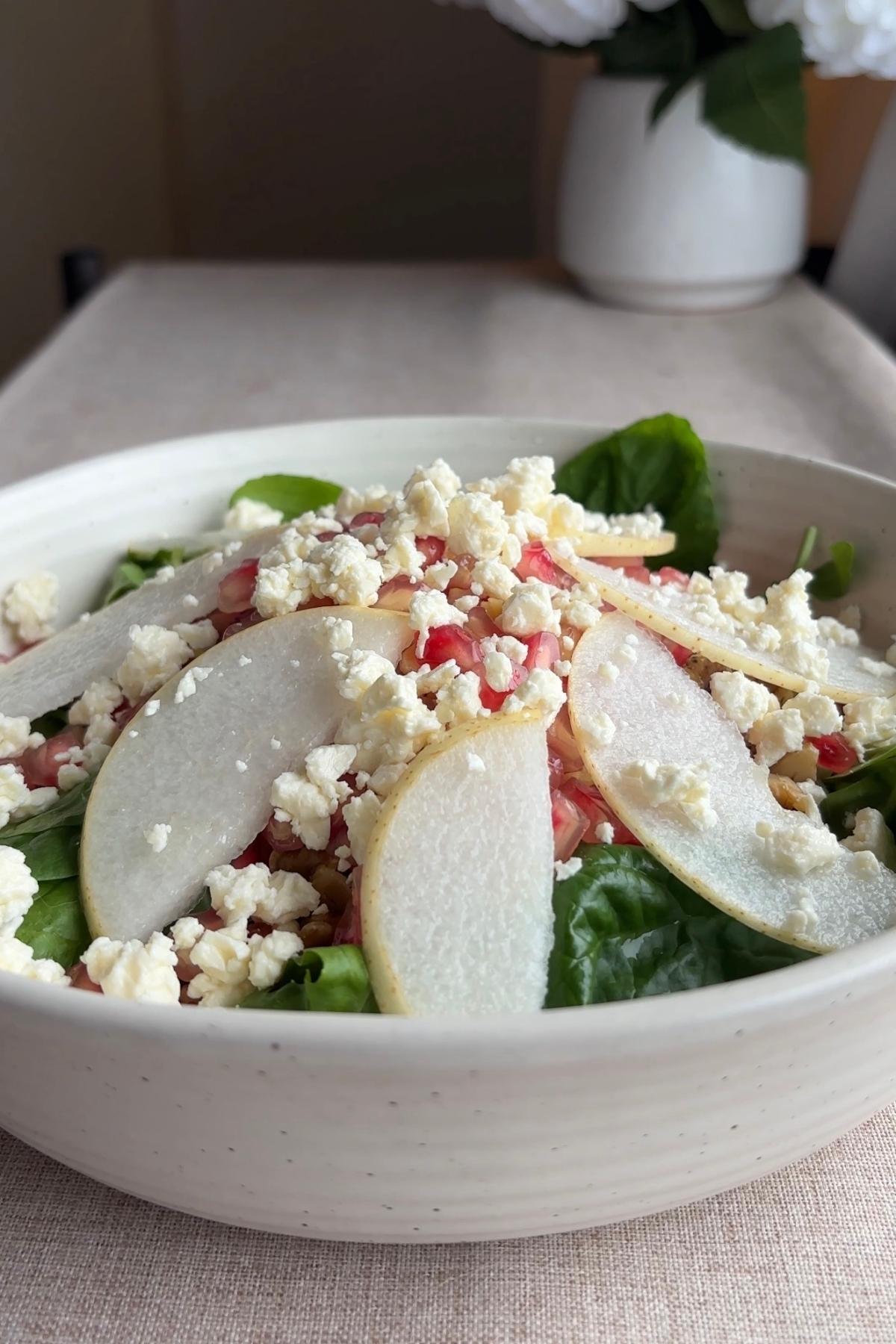 Thinly sliced pears layered over leafy greens in a bowl, with crumbled cheese and pomegranate arils added on top.