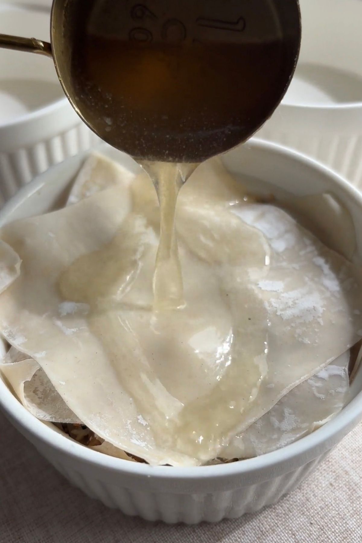 Hot broth being poured over layered wonton wrappers and filling in a ramekin.
