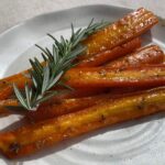 Glossy roasted carrots arranged on a plate with a fresh rosemary sprig on top.
