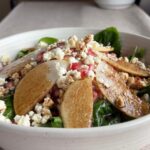 Finished pear salad in a shallow white bowl with spinach, arugula, sliced pears, pomegranate arils, chopped nuts, and crumbled cheese on a neutral linen surface.