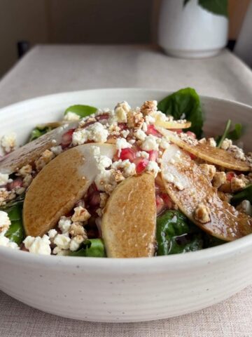 Finished pear salad in a shallow white bowl with spinach, arugula, sliced pears, pomegranate arils, chopped nuts, and crumbled cheese on a neutral linen surface.