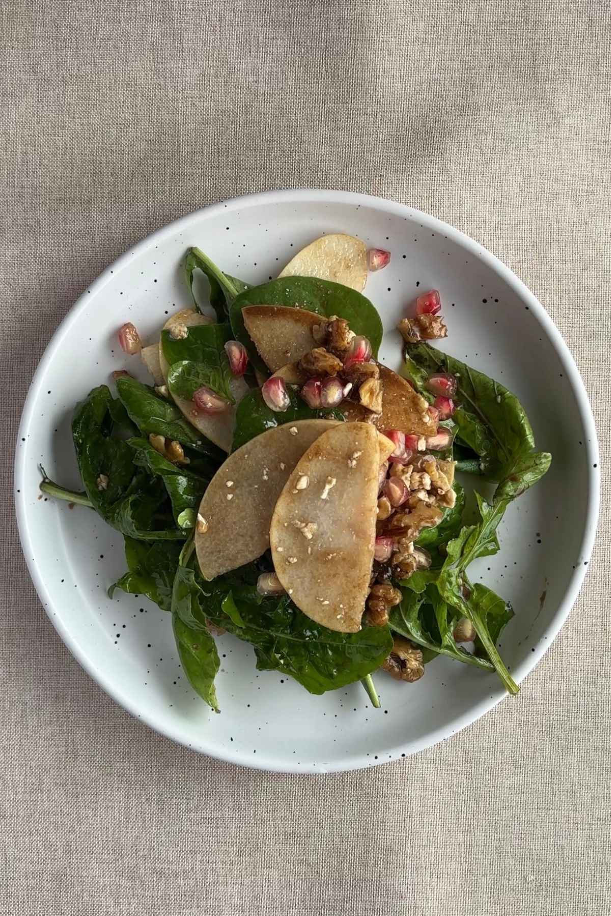 Overhead view of pear salad arranged in a white bowl with leafy greens, thin pear slices, pomegranate arils, and chopped nuts.