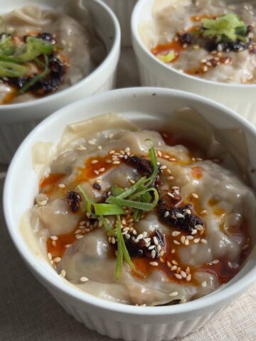 Steamed wonton dumpling bake in a ramekin topped with chili oil, sesame seeds, and scallions.