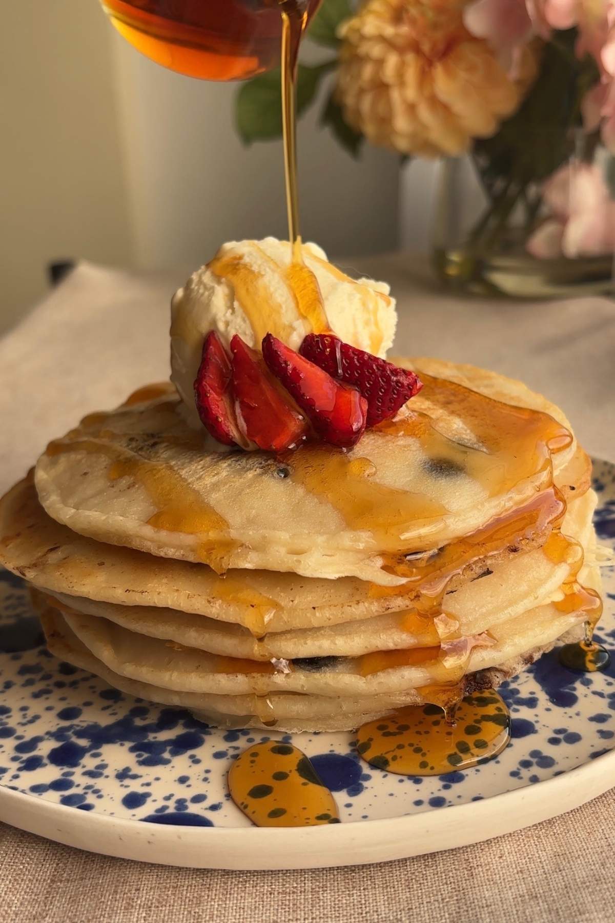 Syrup being poured over pancakes topped with strawberries and vanilla ice cream.