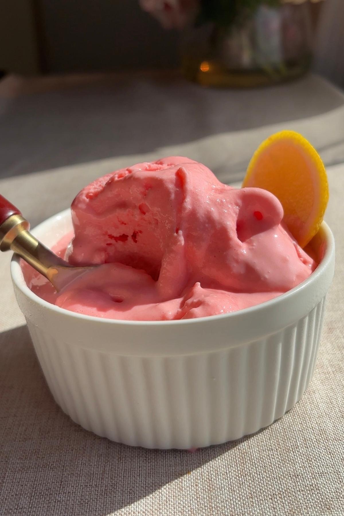 Pink sorbet scoops melting slightly in a white ramekin, with a spoon digging into the center.