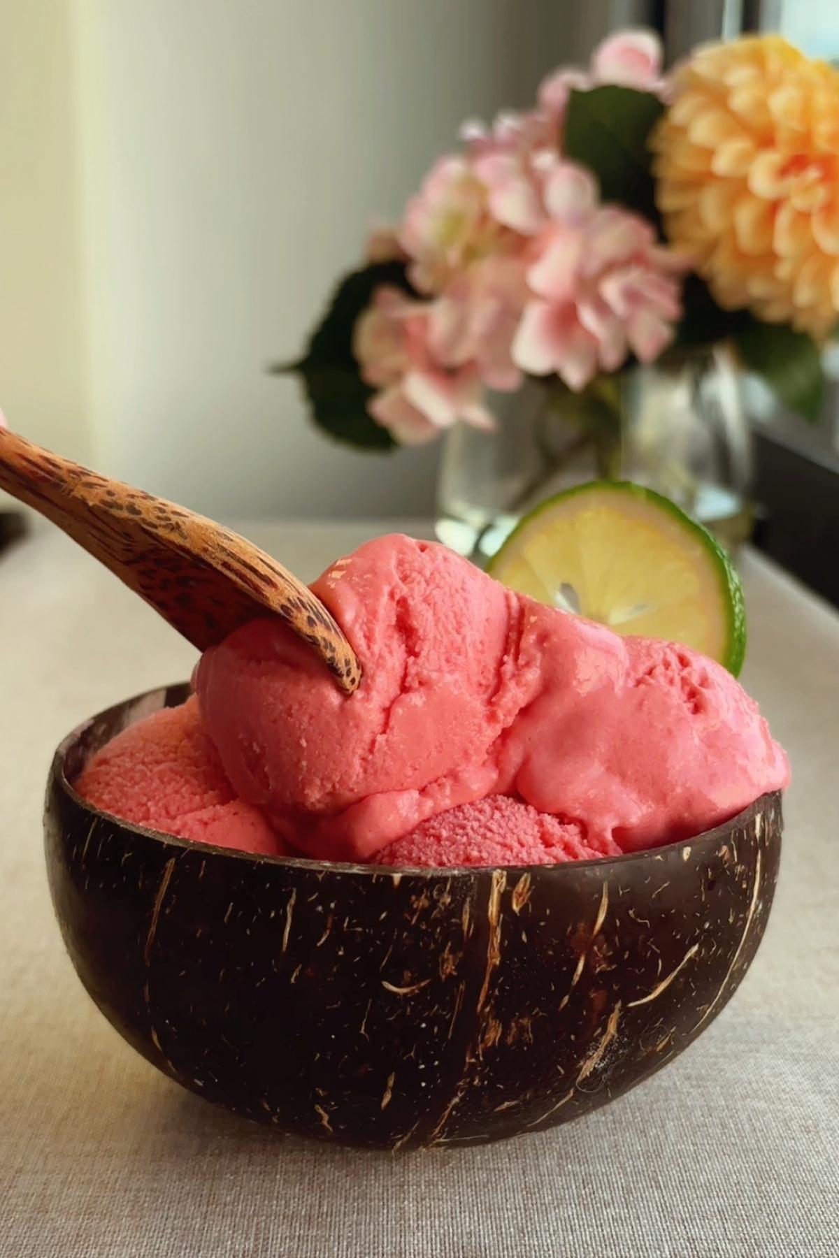 Pink sorbet scooped into a coconut bowl with a wooden spoon and lime slice.