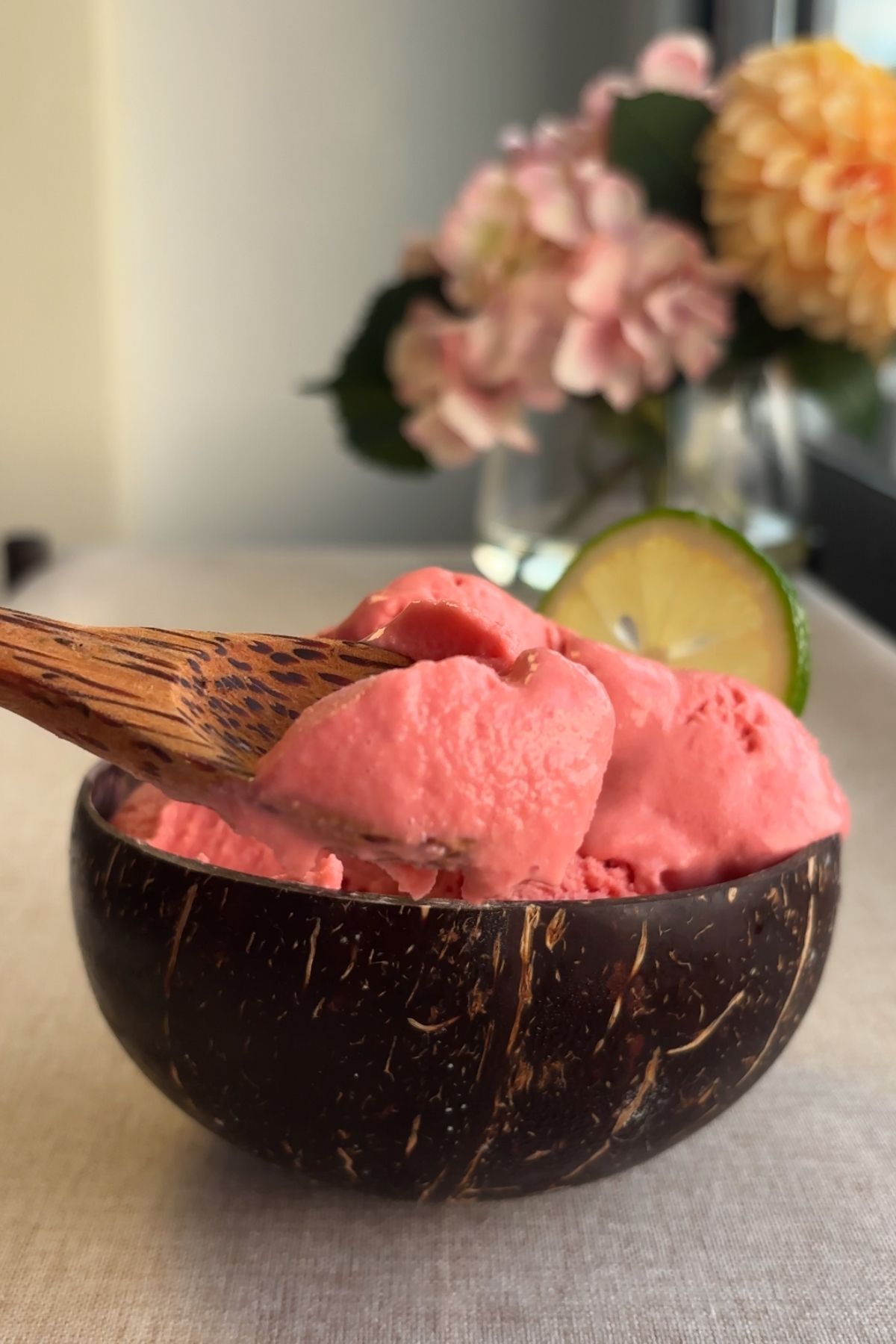 Wooden spoon lifting a scoop of pink sorbet from a coconut bowl.
