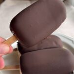 Hand holding a vegan chocolate-coated ice cream bar with smooth snap shell