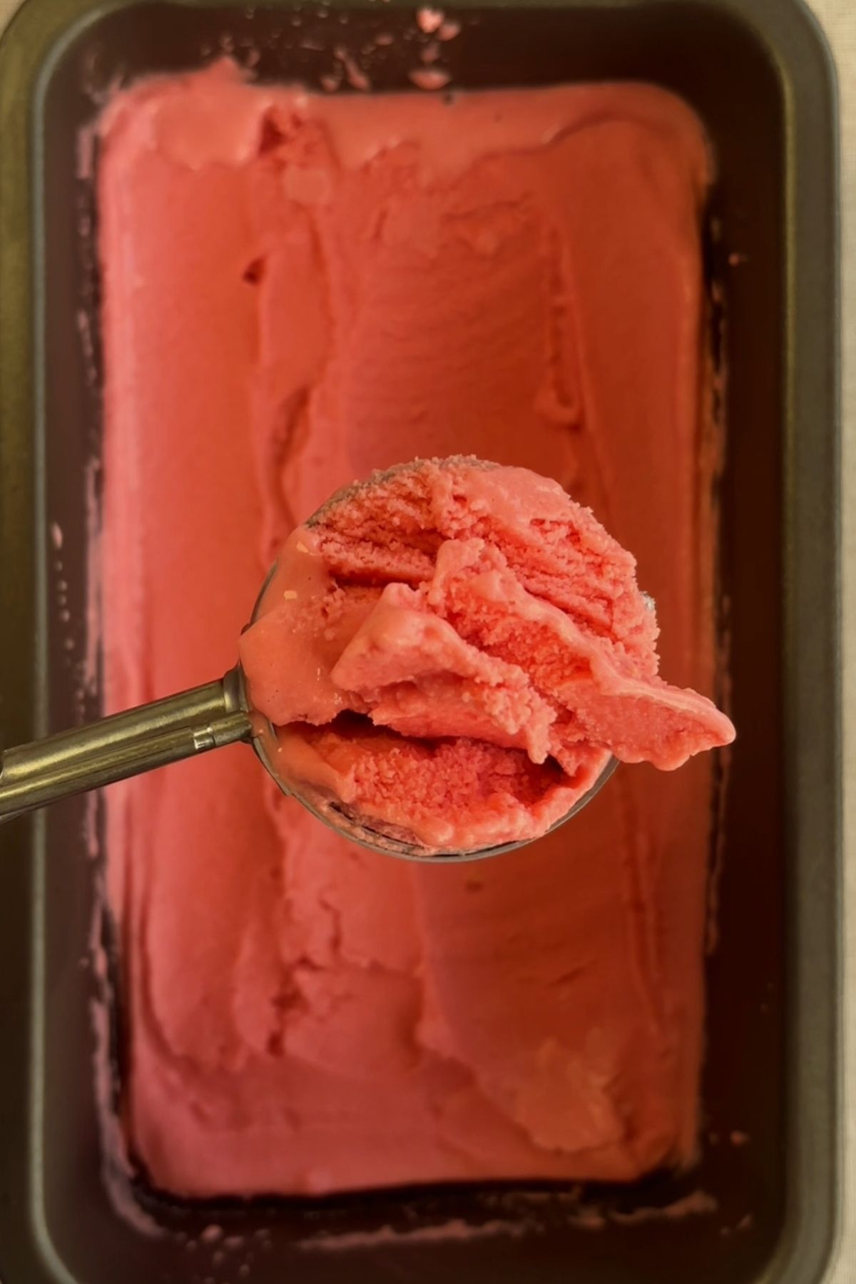 Ice cream scoop lifting pink sorbet from a loaf pan.
