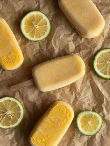 Several mango popsicles on brown paper with fresh lime slices placed between them.