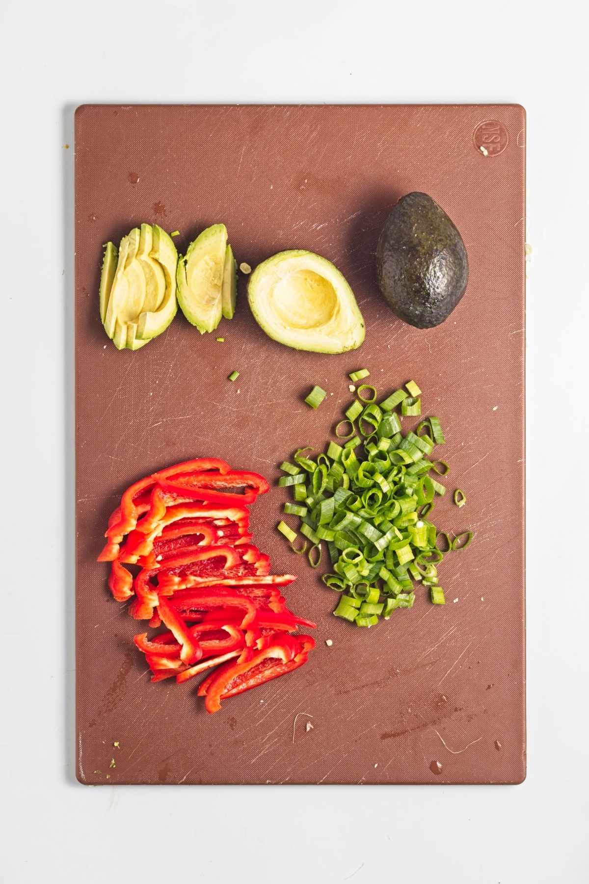 Sliced avocado, red bell pepper, and green onions on a cutting board