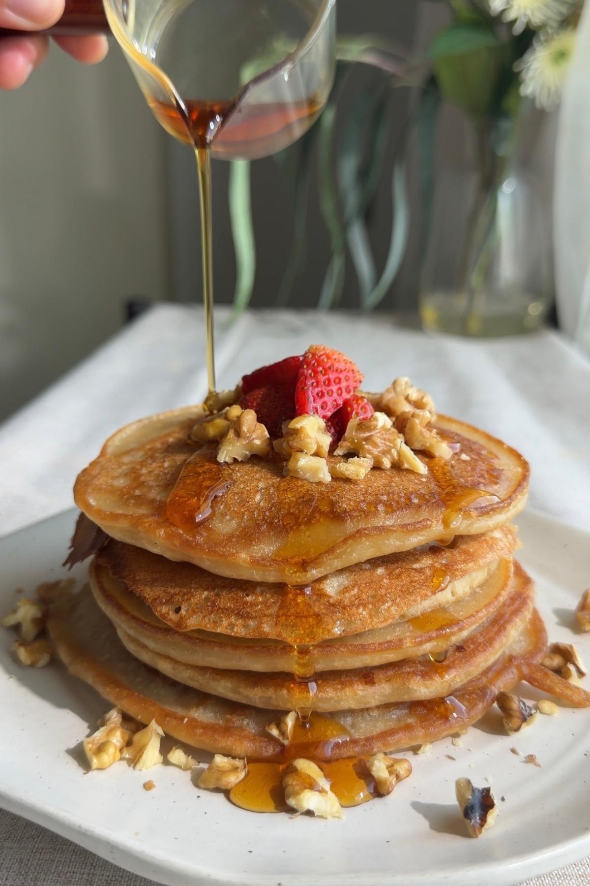 Syrup pouring over a pancake stack with strawberries and chopped nuts.