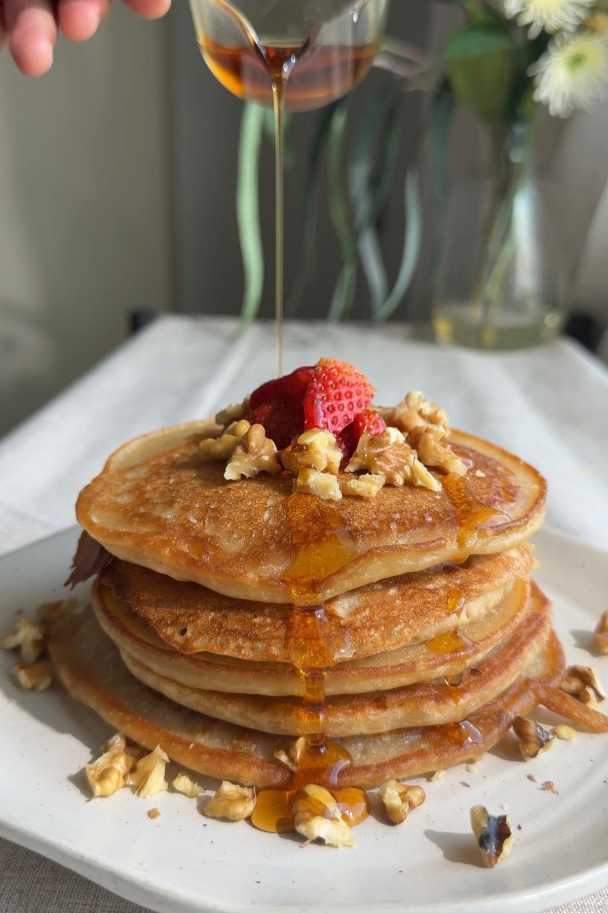 Syrup pouring onto a pancake stack topped with strawberries and chopped nuts.