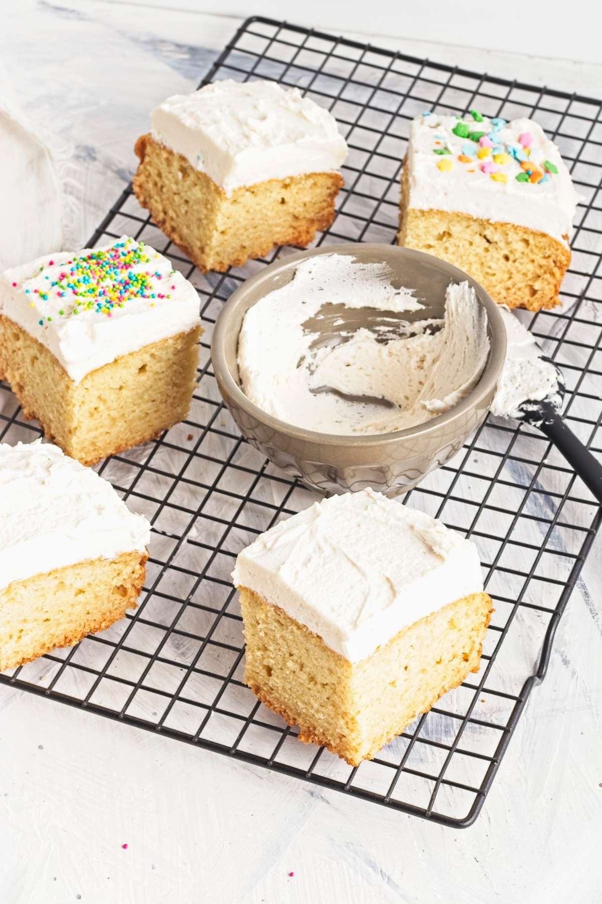 Frosted cake squares on a cooling rack with a bowl of frosting.