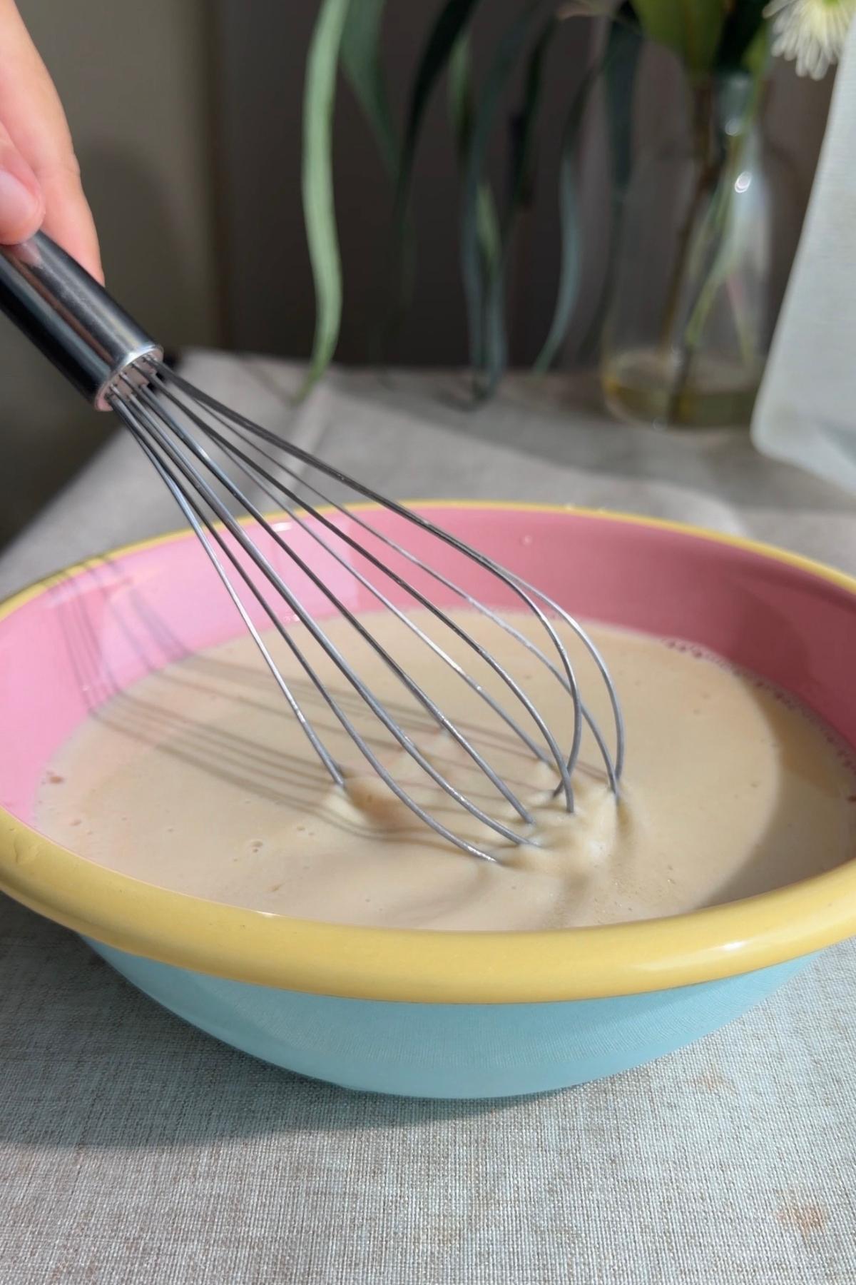 Whisk stirring a pale liquid mixture in a colorful mixing bowl.
