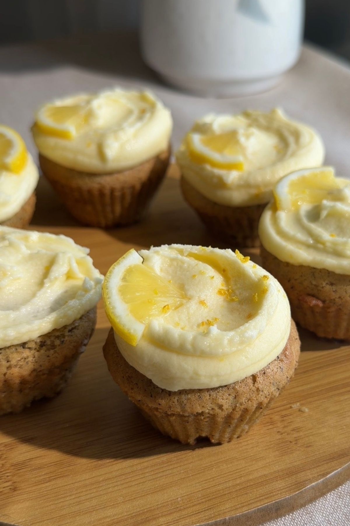 Frosted cupcakes topped with lemon slices and lemon zest on a wooden board.