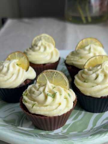 Five frosted ginger lime cupcakes garnished with lemon slices on a patterned serving plate.