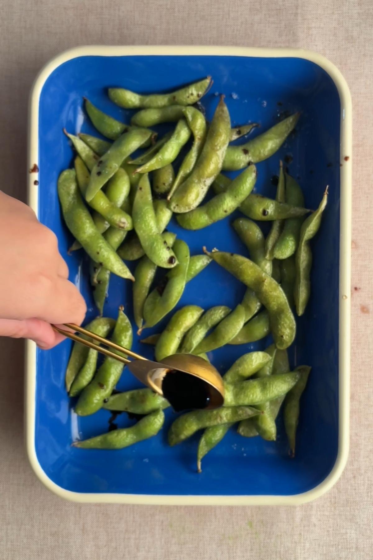 Edamame pods in a baking dish with soy sauce being drizzled over the top.