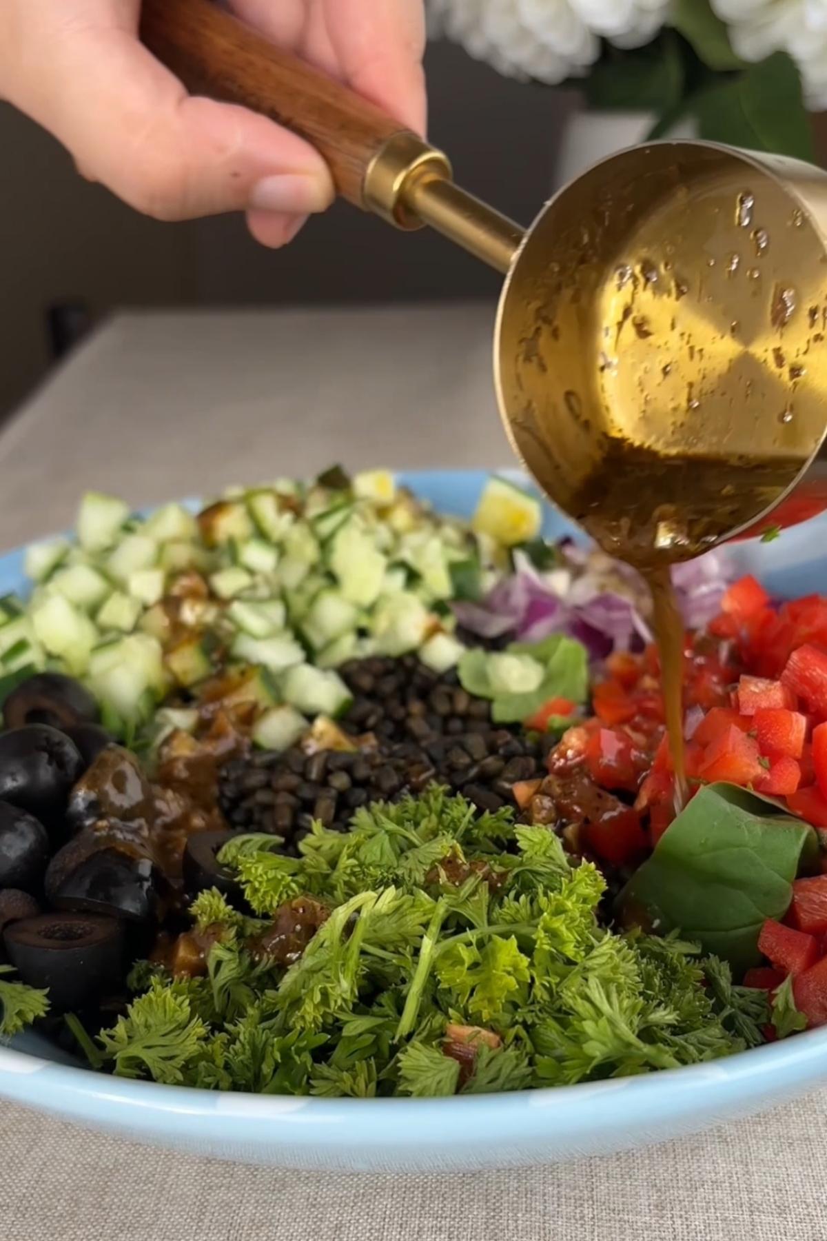 Warm dressing being poured over lentils, vegetables, olives, and parsley in a bowl.