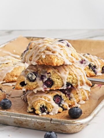Stack of glazed blueberry scones on a baking sheet with fresh blueberries.