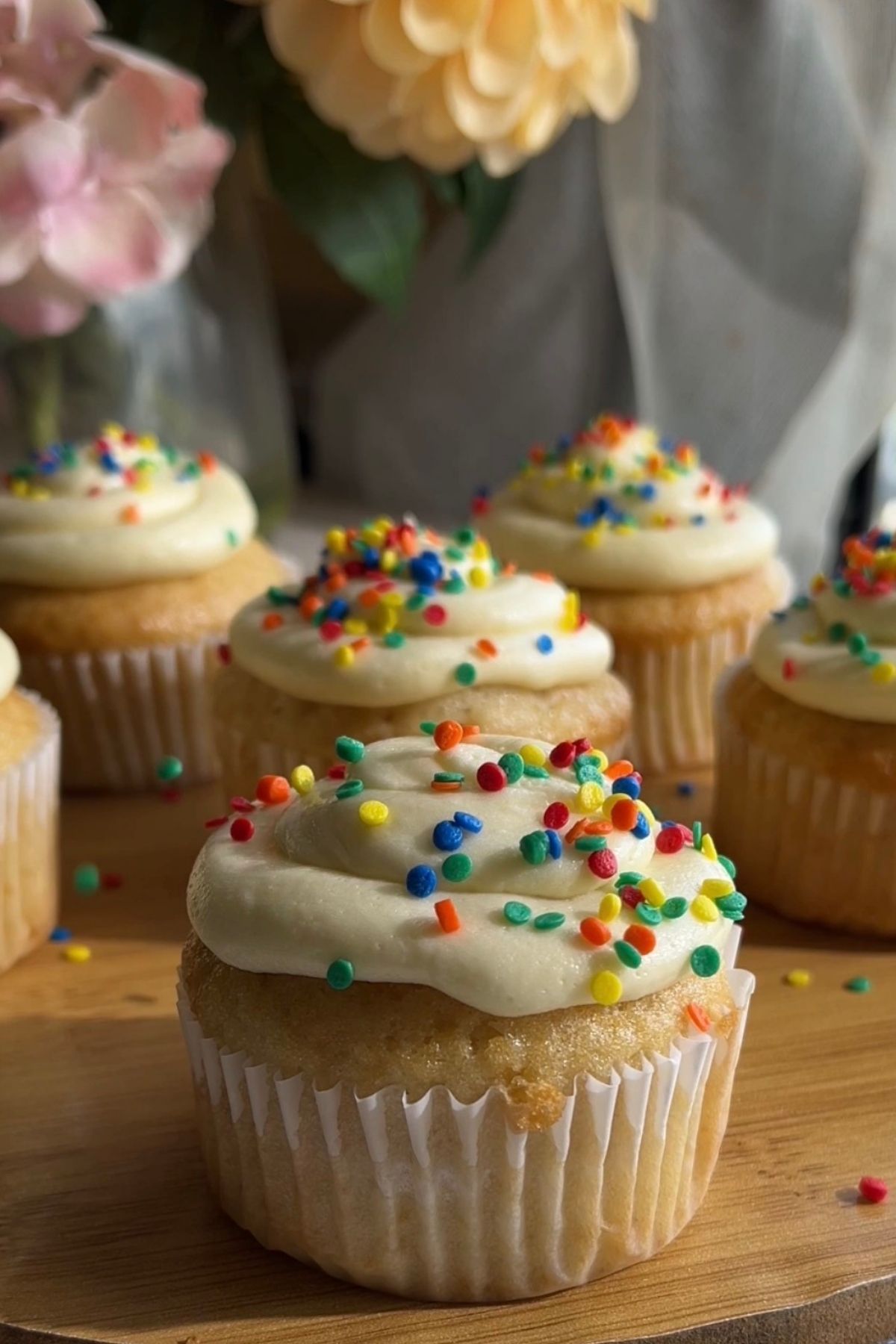 A frosted cupcake with rainbow sprinkles on a wooden board.