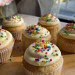 Frosted cupcakes with rainbow sprinkles on a wooden board.