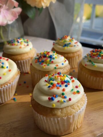 Frosted cupcakes with rainbow sprinkles on a wooden board.