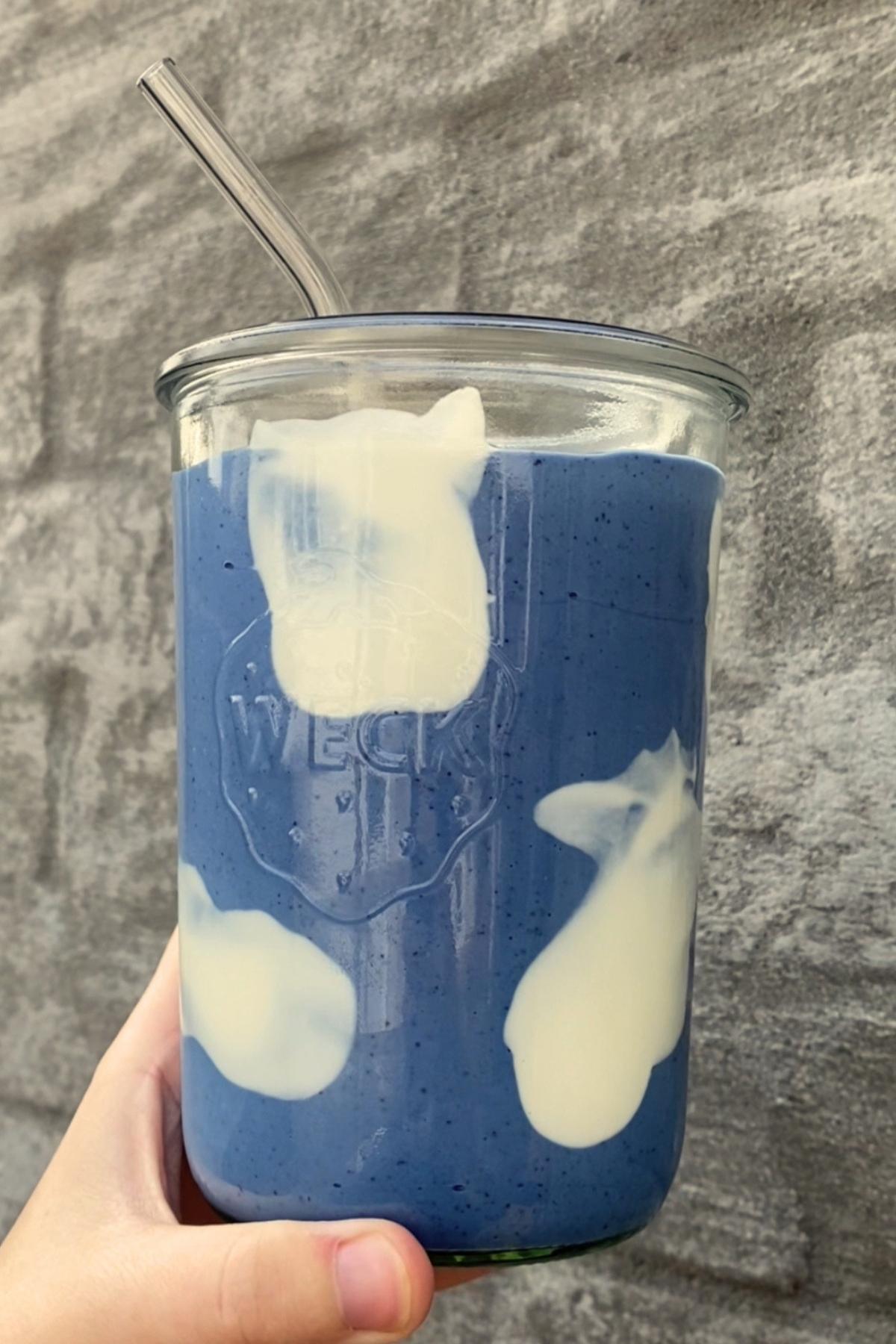 Hand holding a glass jar of blue spirulina smoothie with creamy yogurt swirls and a reusable glass straw.