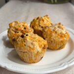 Four vegan carrot cake muffins on a white plate with shredded carrot, oats, and dried cranberries.