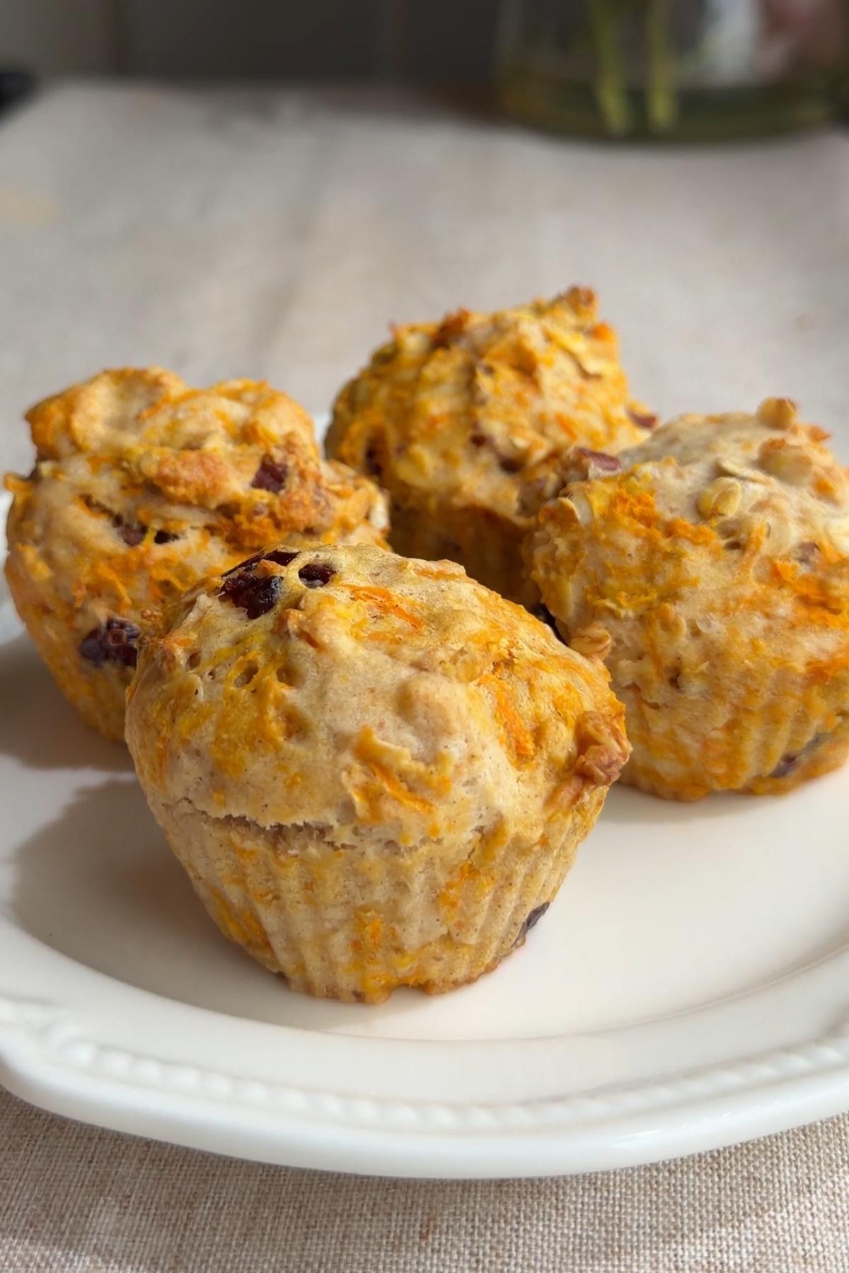Four vegan carrot cake muffins on a white plate with shredded carrot, oats, and dried cranberries.