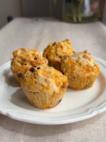 Four vegan carrot cake muffins on a white plate with shredded carrot, oats, and dried cranberries.