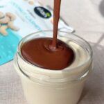 Chocolate sauce being poured over creamy cashew mousse in a glass jar.