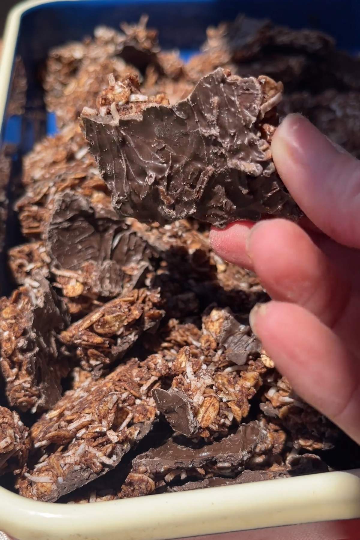 Hand holding a large chocolate cluster from a tray of chocolate coconut granola.