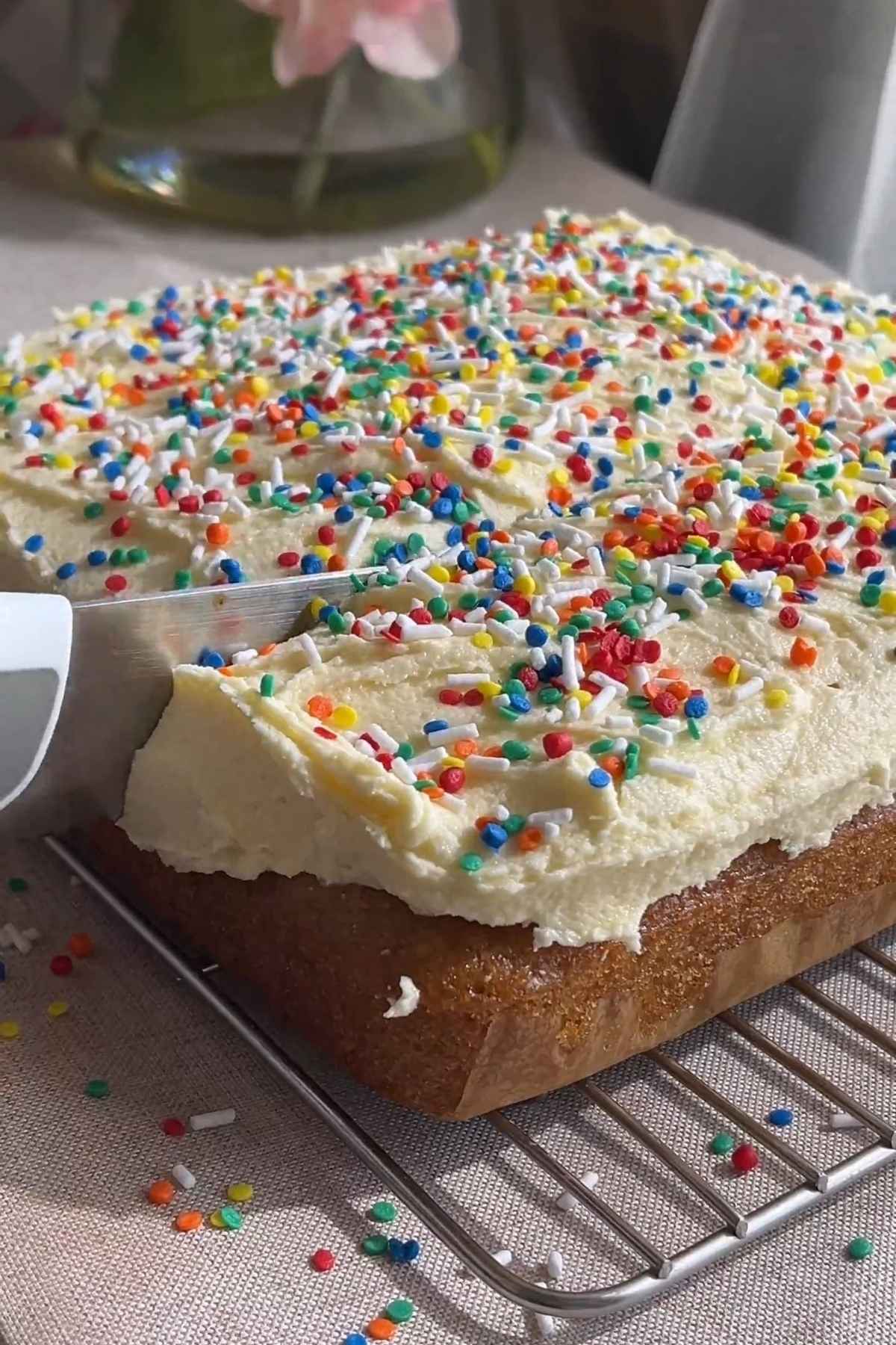 Knife slicing frosted vegan vanilla cake topped with colorful sprinkles on a cooling rack.