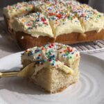 Slice of vegan vanilla cake with vanilla frosting and colorful sprinkles on a white plate.