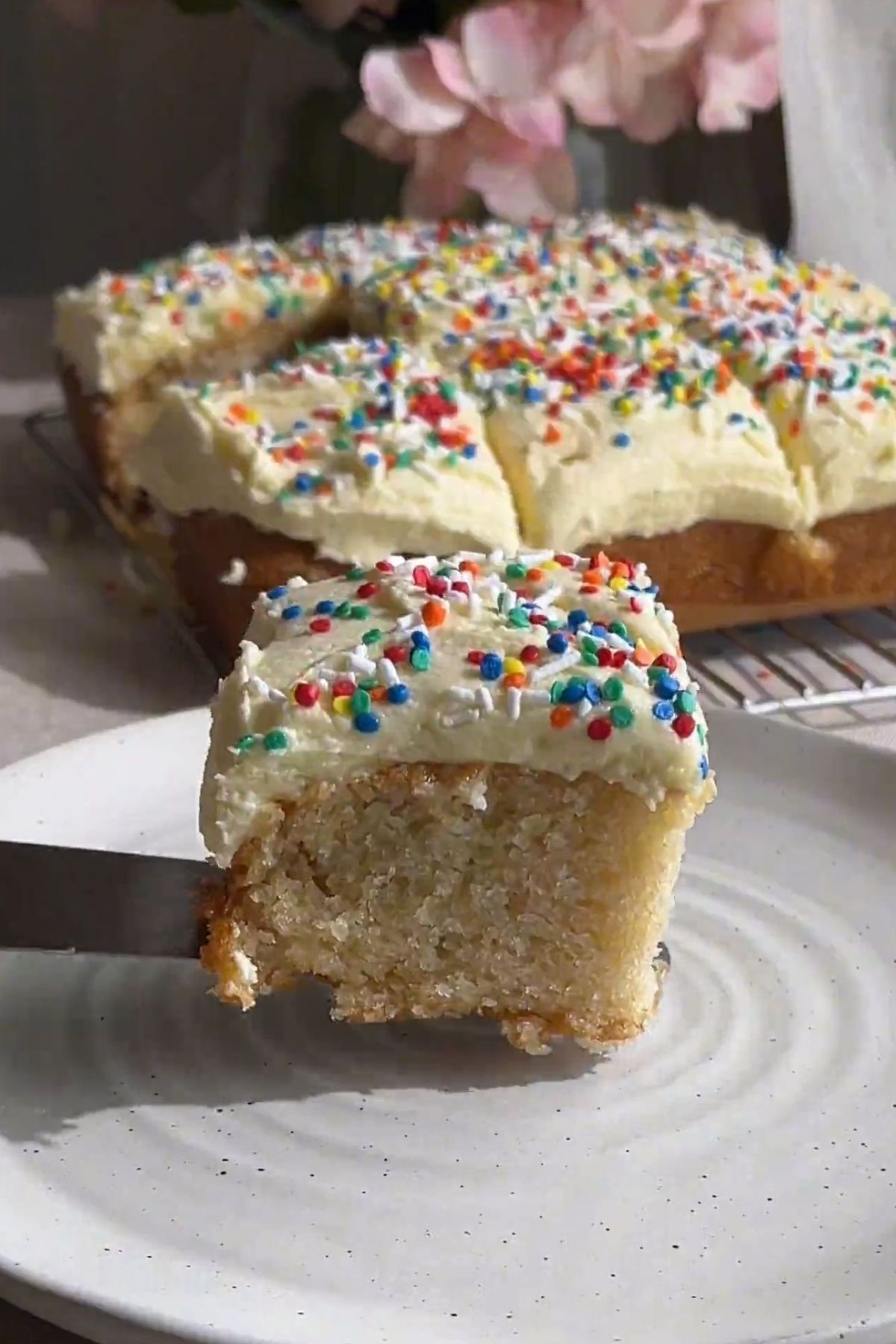 Frosted vegan vanilla cake topped with colorful sprinkles, cut into squares on a cooling rack.
