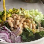 Golden dressing being poured over kale, white beans, avocado, cucumber, and red onion in a salad bowl.