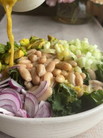 Golden dressing being poured over kale, white beans, avocado, cucumber, and red onion in a salad bowl.