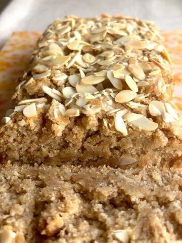 Gluten-free lemon loaf topped with toasted sliced almonds on parchment paper, with one slice cut.