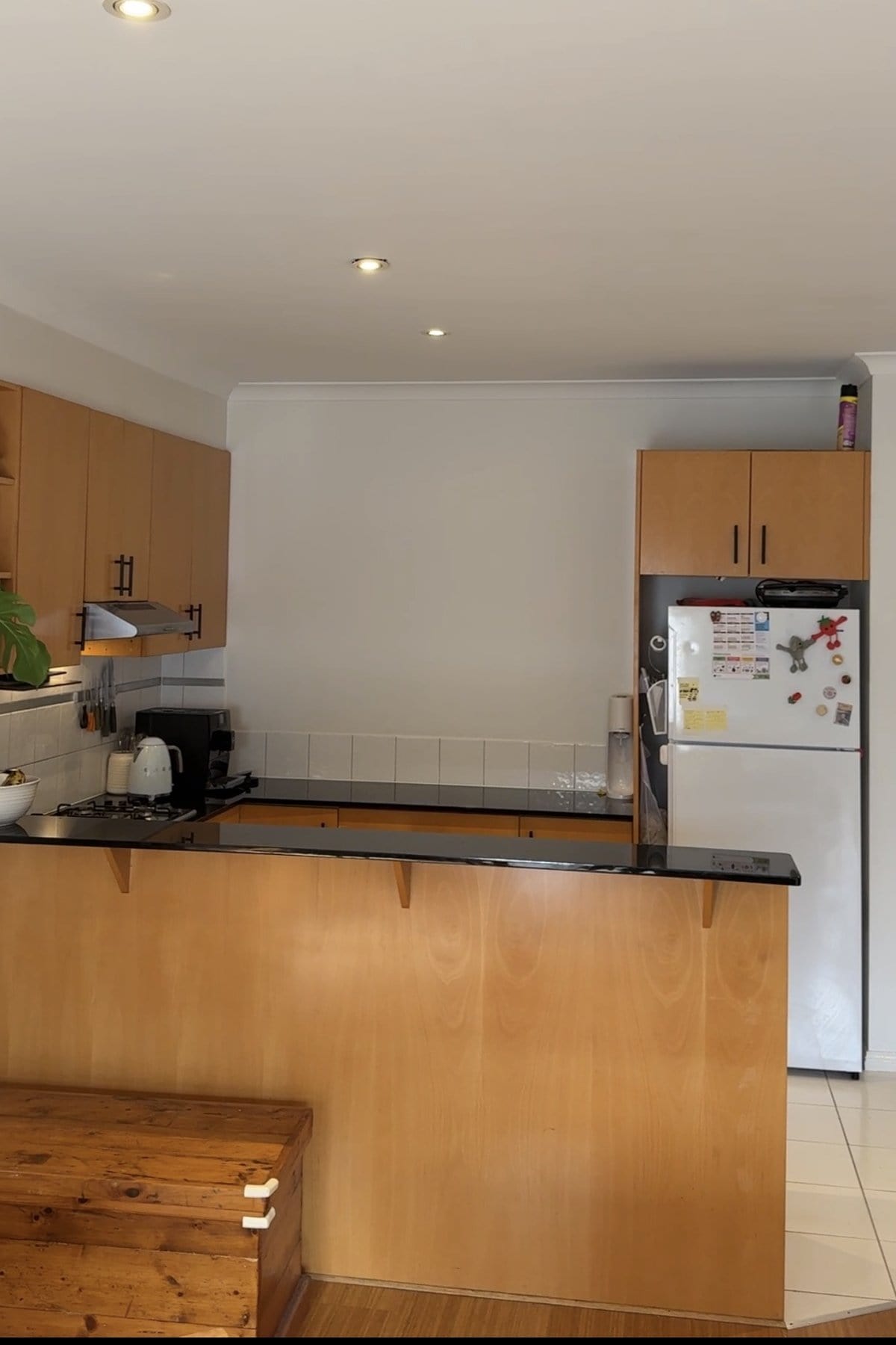 Timber kitchen with a black stone benchtop, breakfast bar and a white fridge.