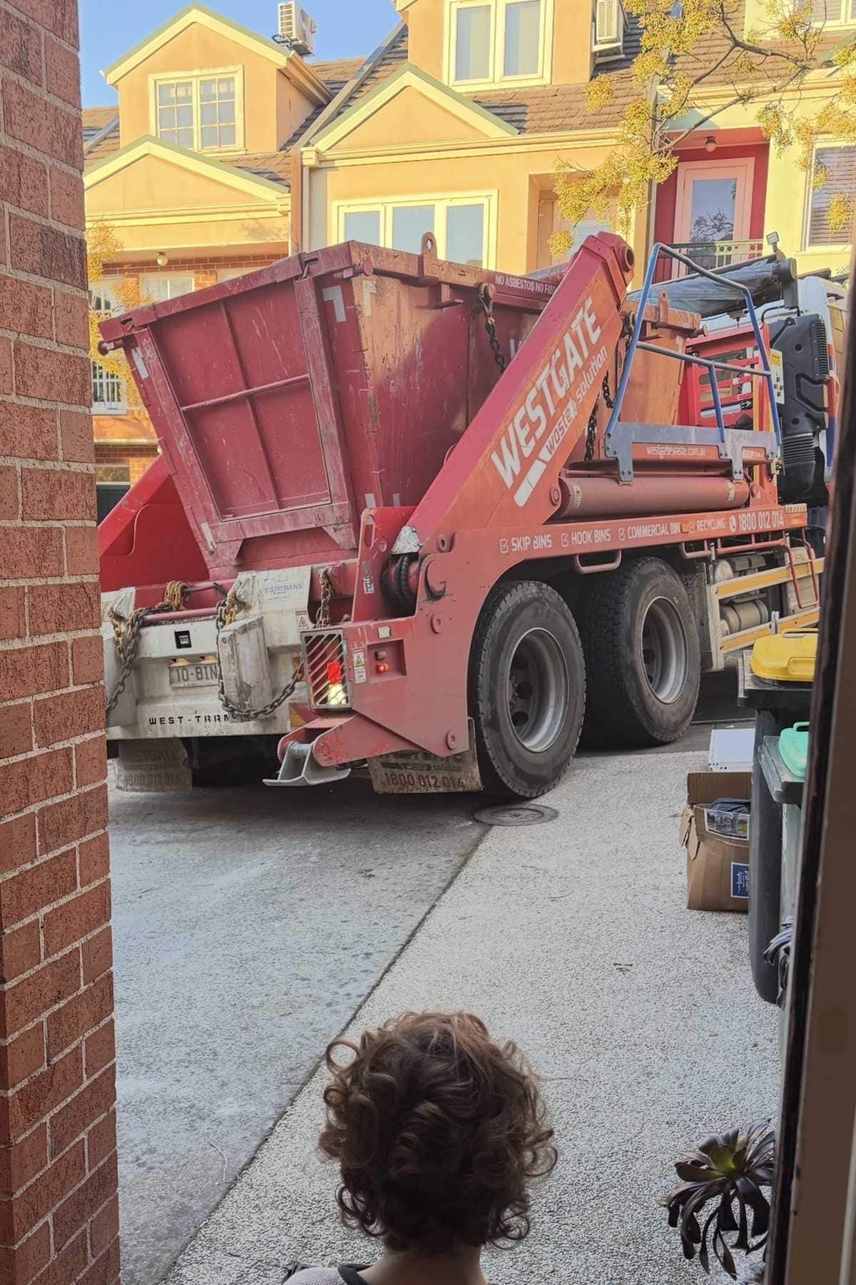 Child watching a skip bin truck in a driveway, seen from a doorway.