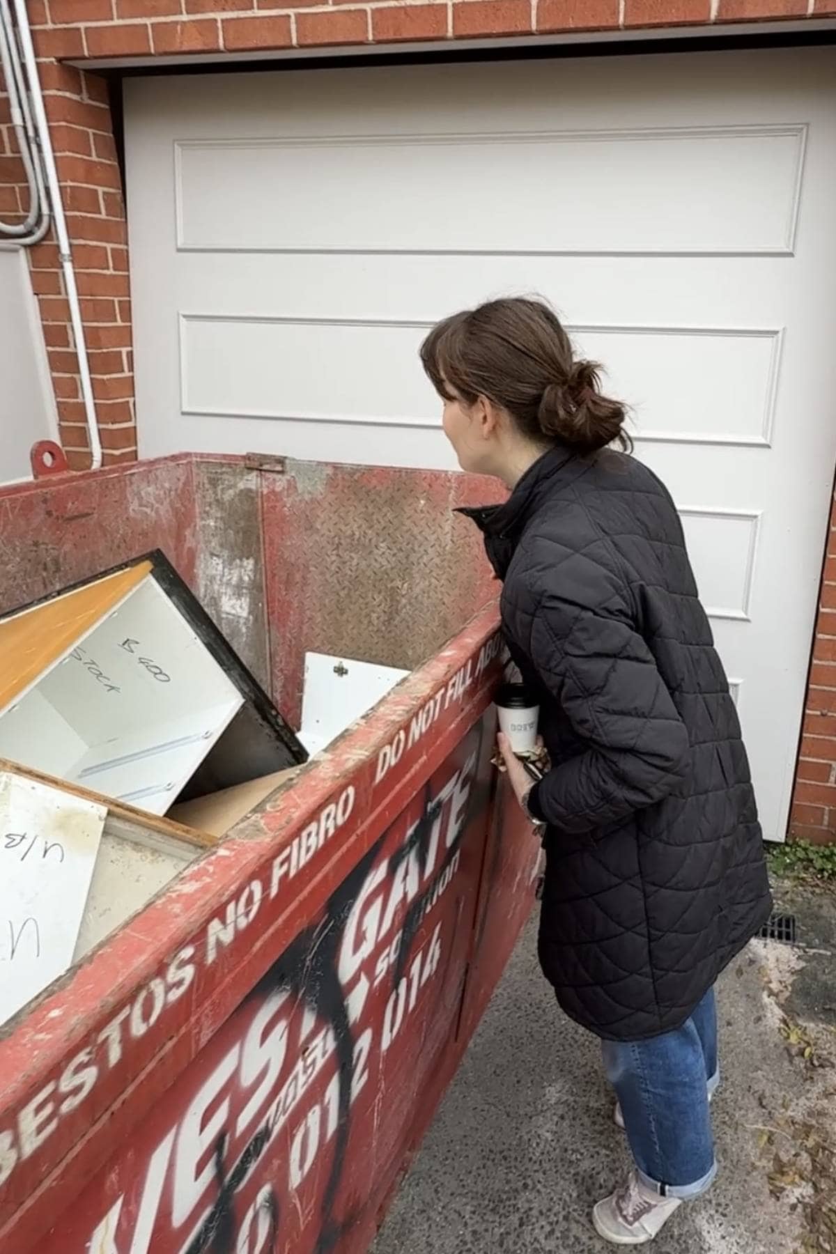Woman in a black coat holding a coffee, looking into a red skip bin outside a garage.