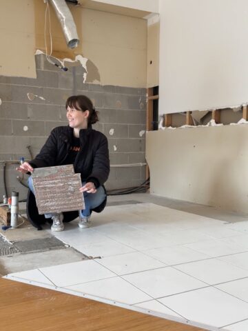 Woman crouching in a gutted room, holding up a floor tile lifted from the slab.