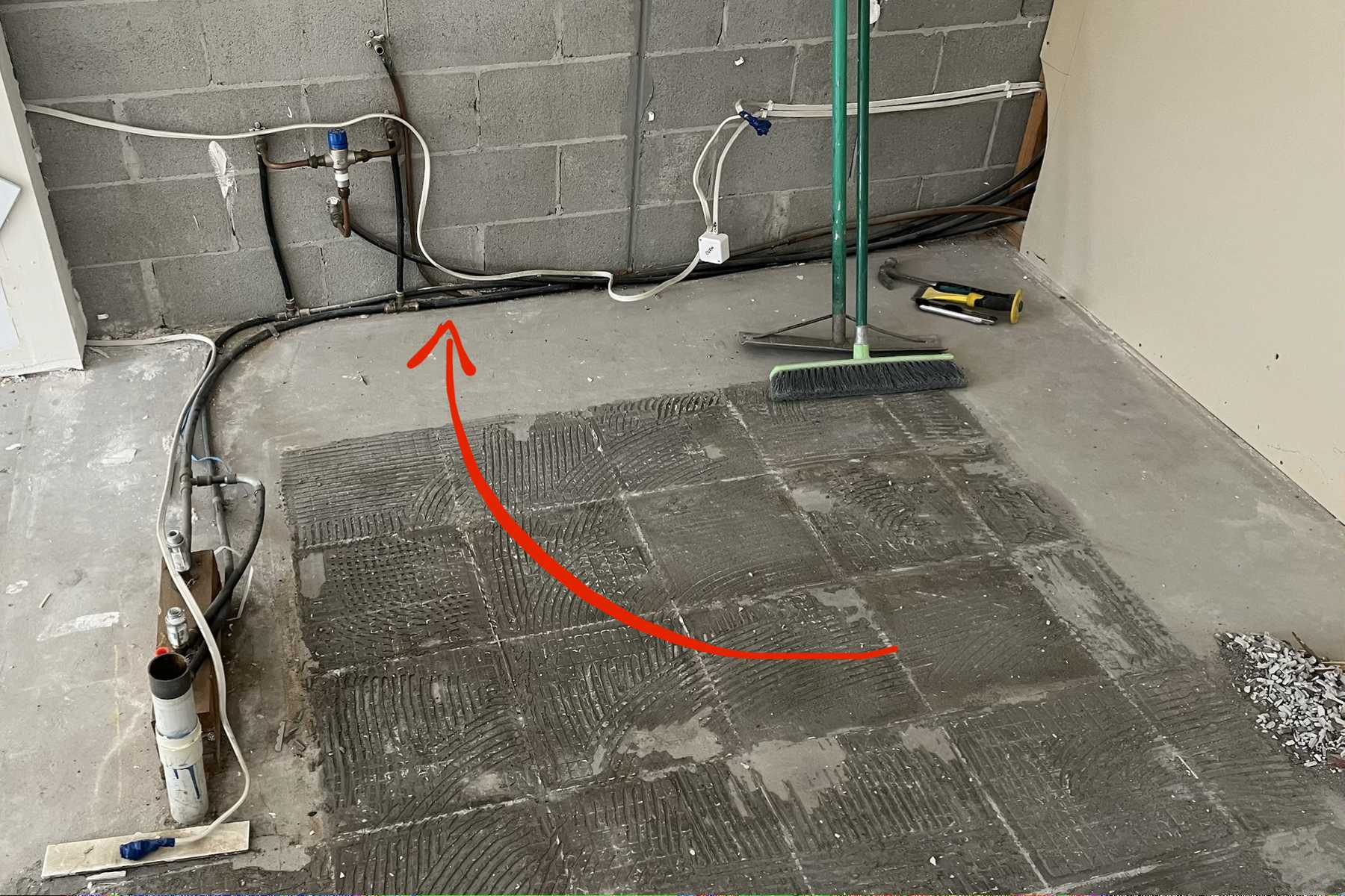 Concrete floor with the tiles lifted, exposed pipework along a block wall and a red arrow marking a spot.
