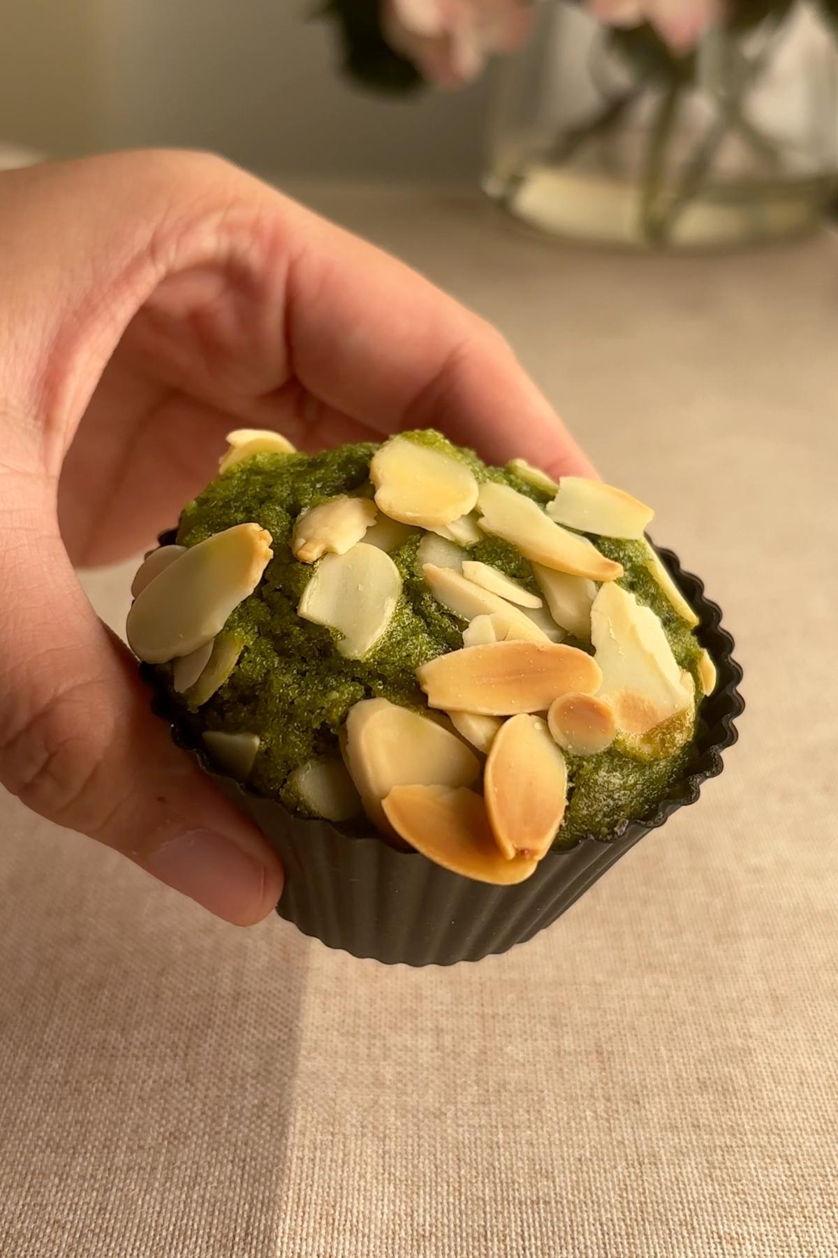 Hand holding a matcha muffin topped with toasted sliced almonds in a black baking cup.