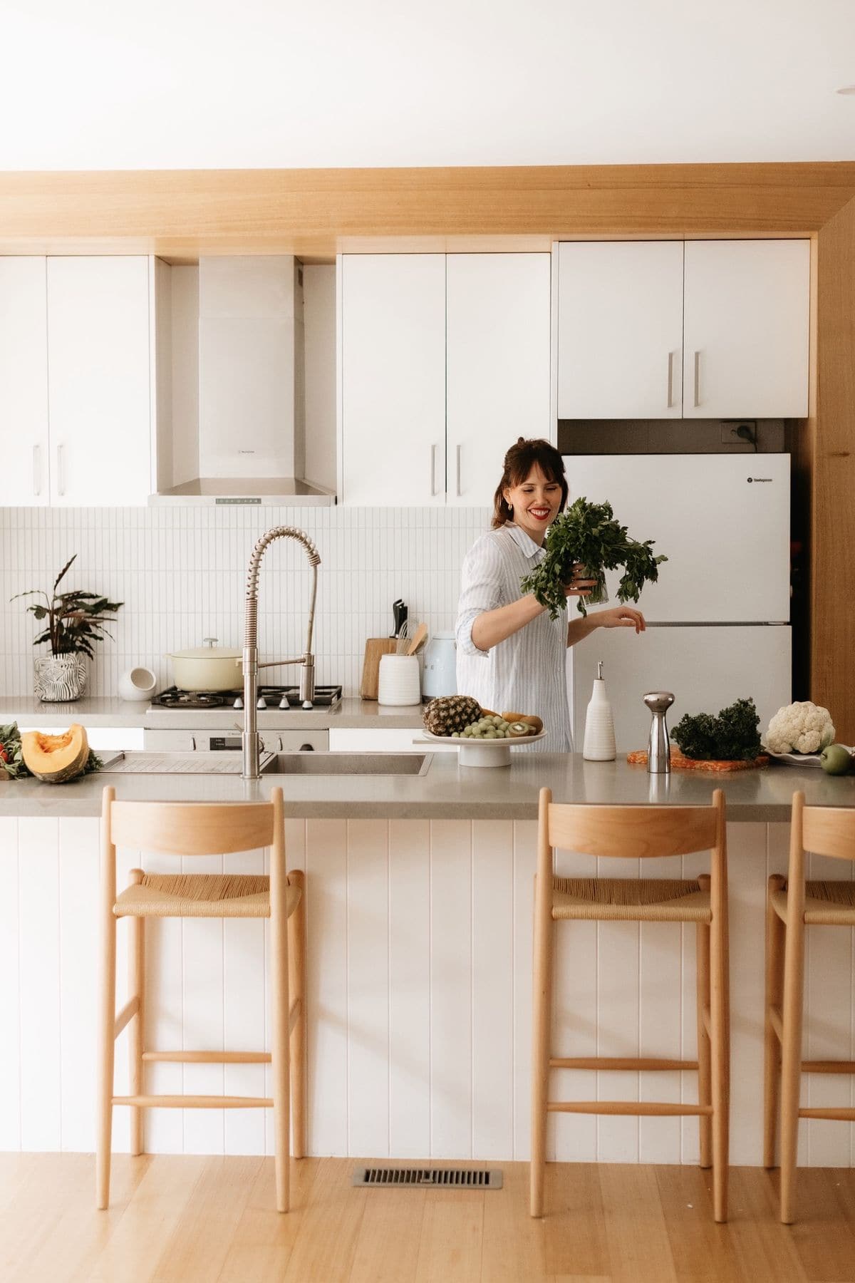 Woman holding a bunch of parsley in a white kitchen with timber trim, a stone island bench and rattan stools.