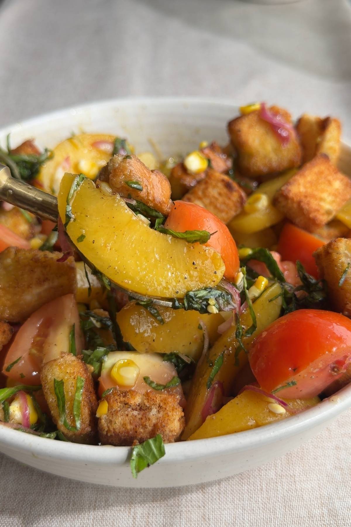 Spoon lifting a peach slice from a panzella salad with tomatoes and croutons.