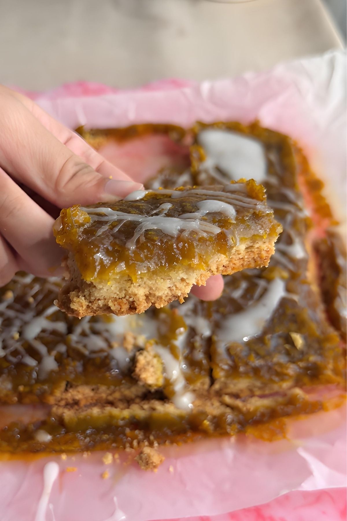 Hand holding a pumpkin pie bar with a thick spiced pumpkin layer, oat crust, and white glaze drizzle.
