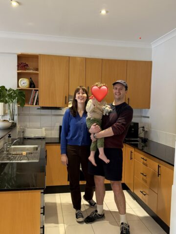 Two adults and a toddler standing in a kitchen with timber cabinets, black benchtops and white tiled floor.
